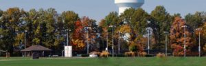 Park with green field, pavilion, car near ornamental grasses, and a line of trees in autumn colors beneath a distant water tower